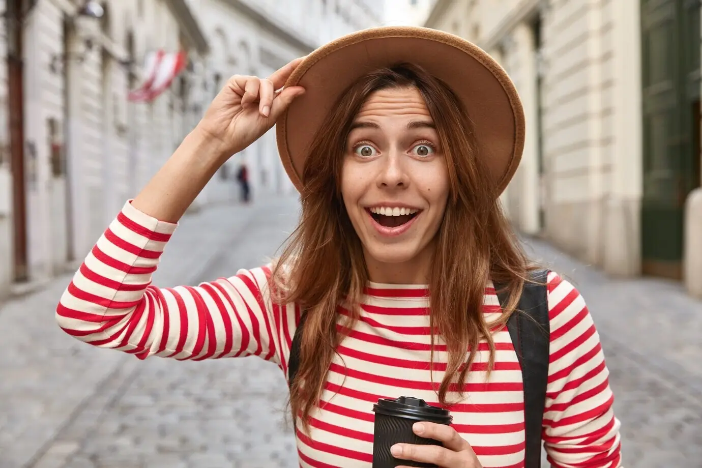 Photo of a surprised, cheerful European woman holding her hat, drinking a takeaway coffee as she walks along a city street.